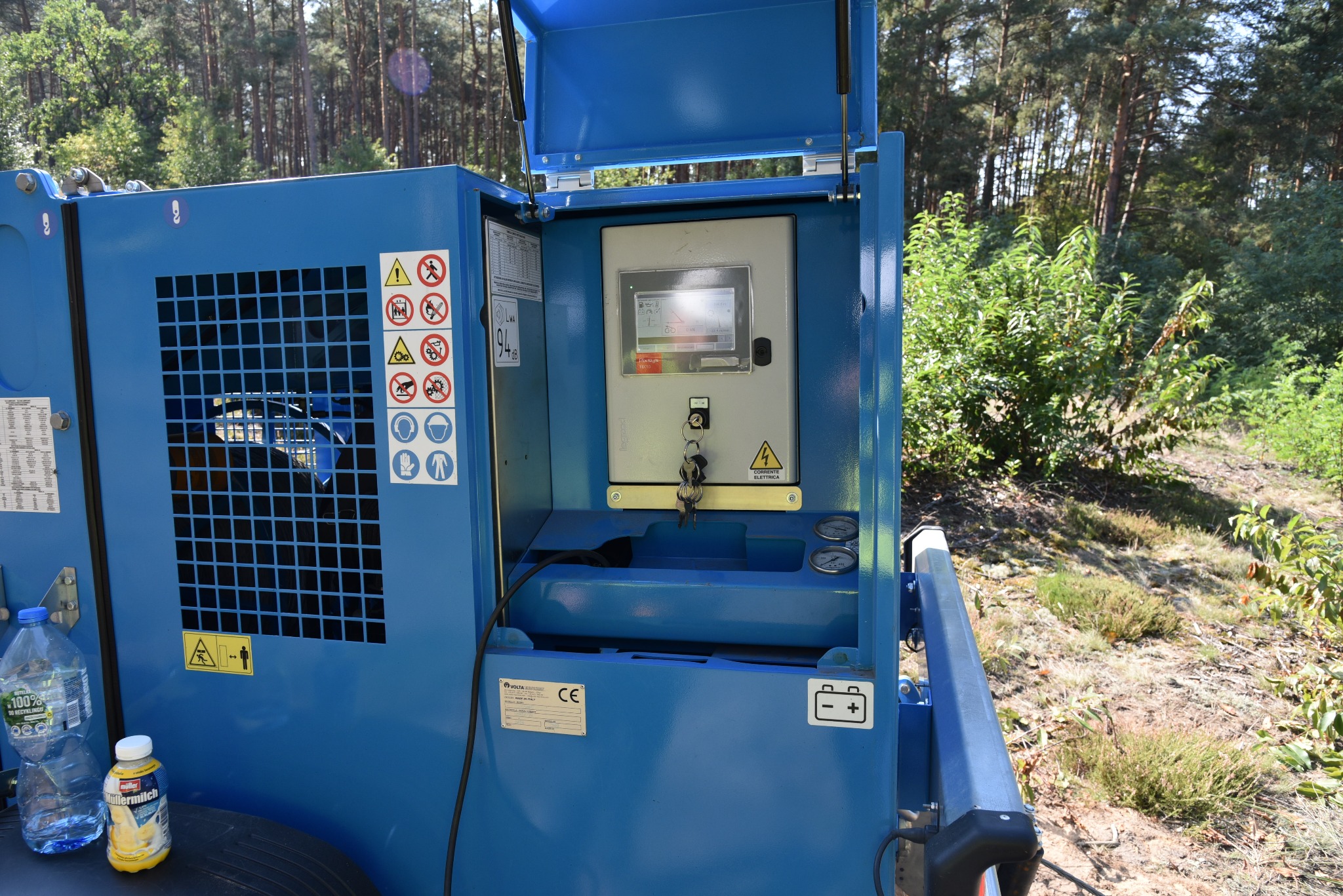 Niebieska, przenośna stacja zasilania na tle leśnym, z otwartą obudową ukazującą panel sterowania z kluczykami; butelki wody i napoju na dolnej półce.