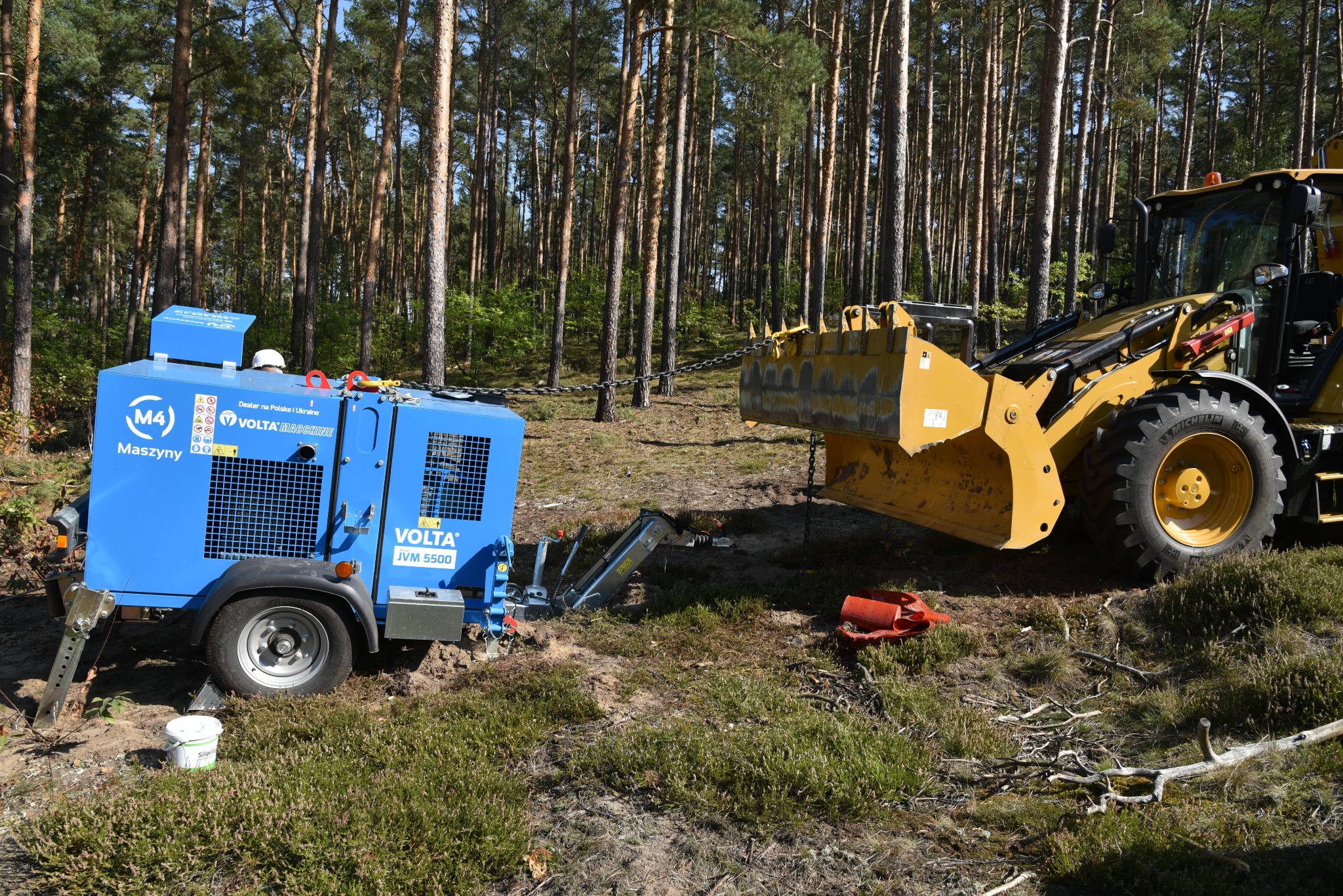 Żółta koparko-ładowarka ciągnie niebieski agregat prądotwórczy Volta JVM 5500 na przyczepie w zalesionym terenie, widoczna łańcuchowa lina holownicza.