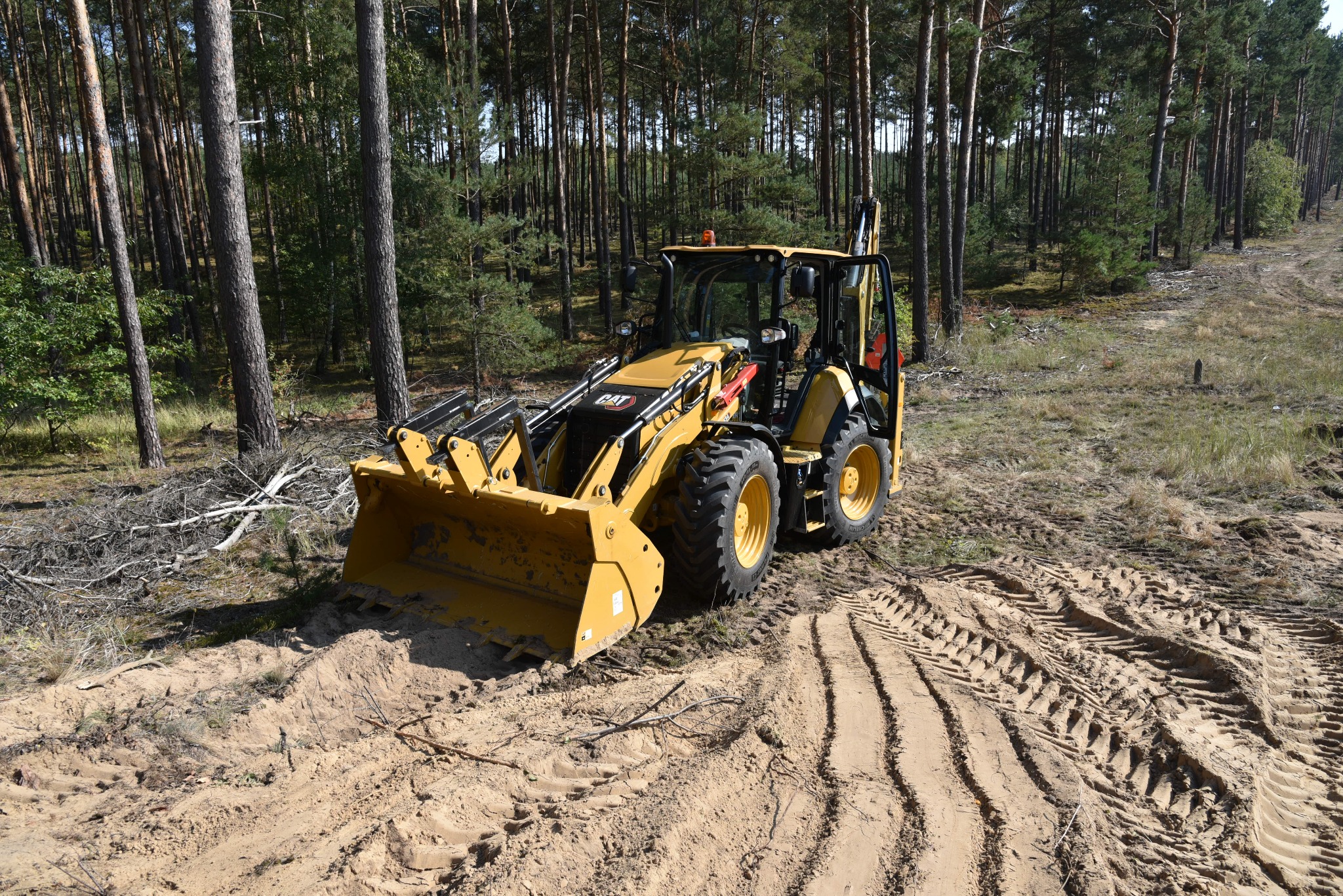 Żółta koparko-ładowarka Caterpillar na piaszczystym terenie leśnym, widoczne ślady opon i przygotowany teren pod wykop.
