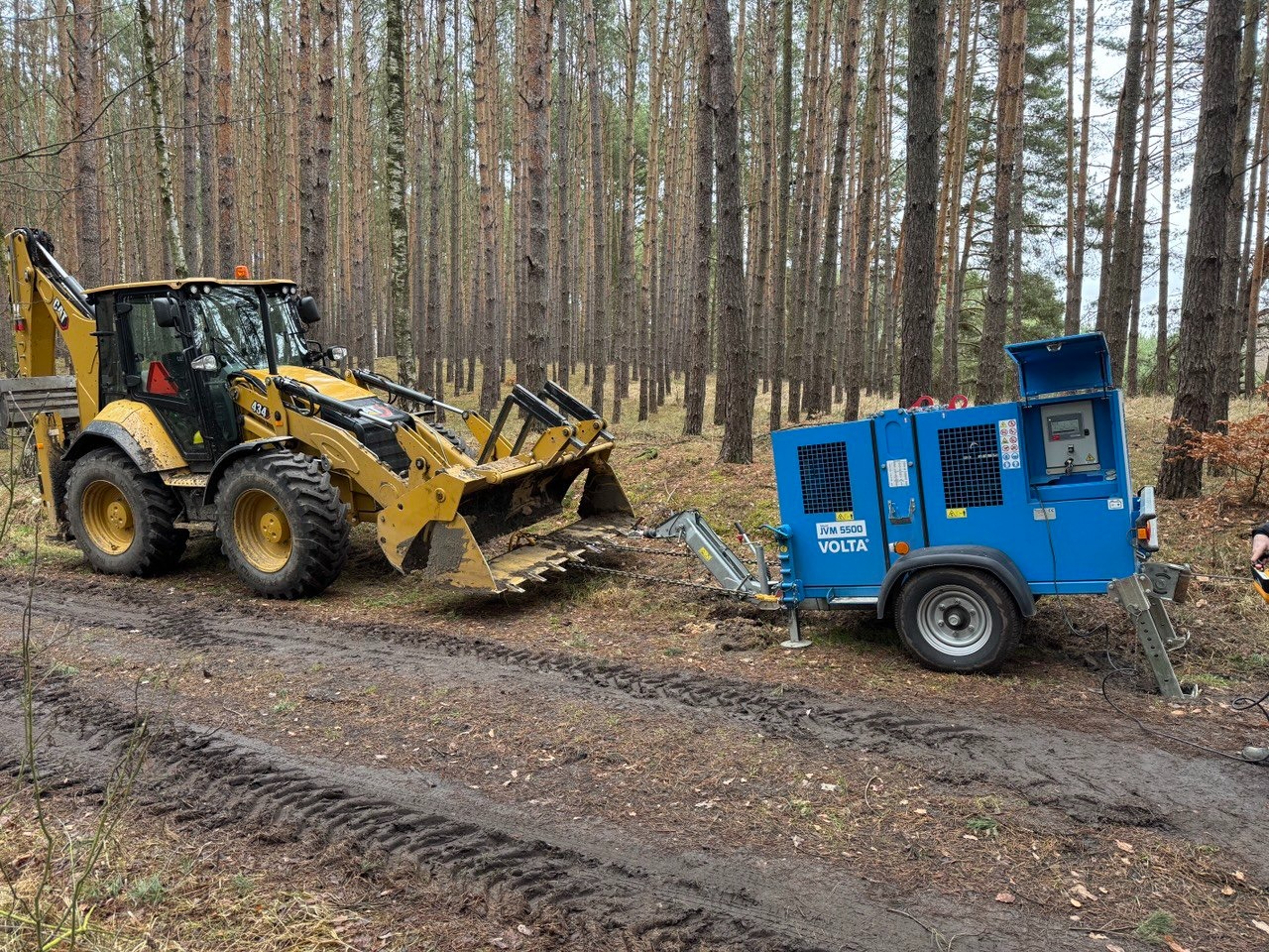Żółta koparko-ładowarka CAT 434 z niebieskim agregatem prądotwórczym Volta IVM 5500 na leśnej drodze z wyraźnymi śladami opon.