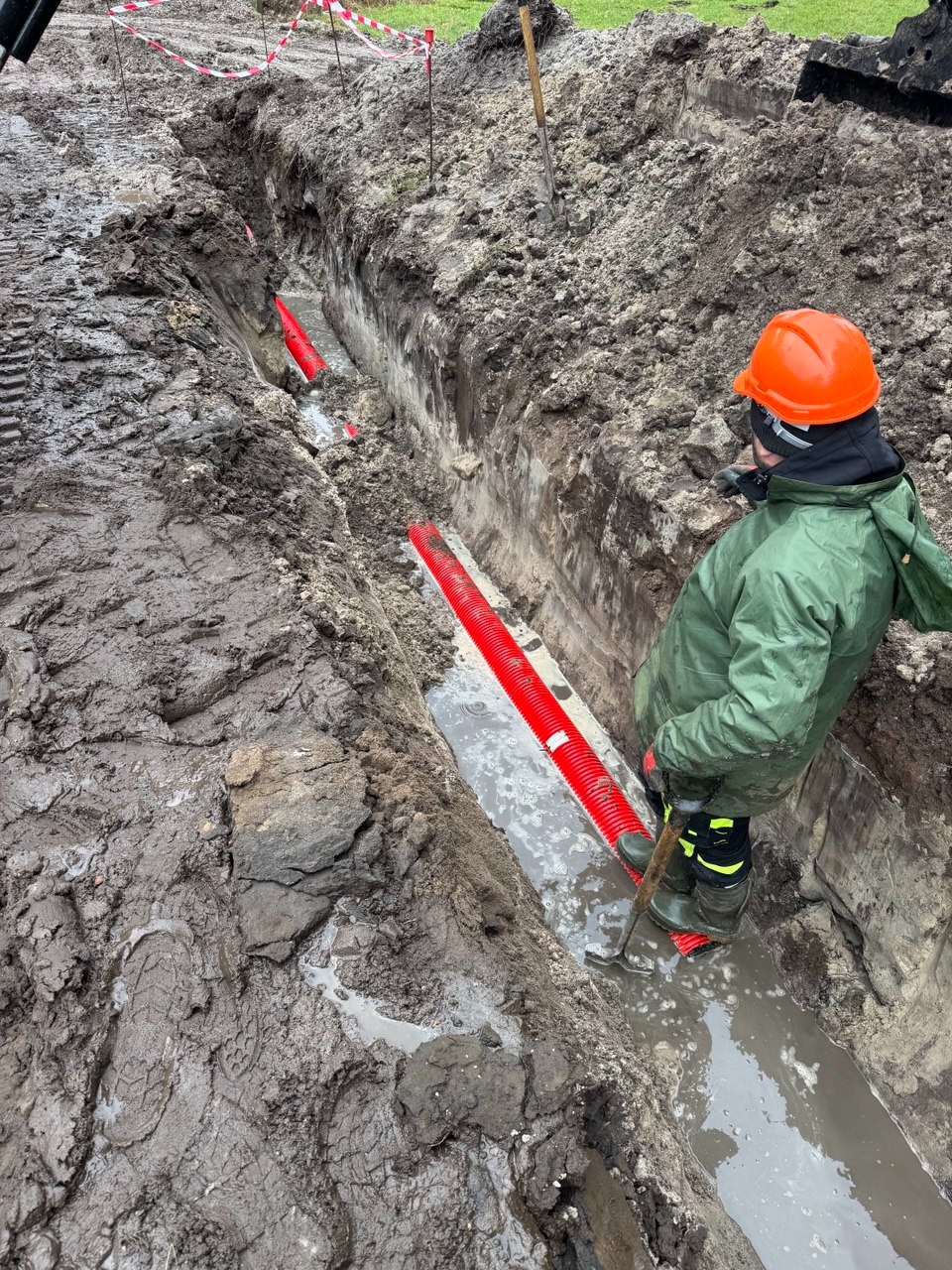 Wykop z ułożonymi czerwonymi rurami karbowanymi, częściowo zalany wodą. Pracownik w kasku i zielonej kurtce nadzoruje ułożenie rur w błotnistym terenie.