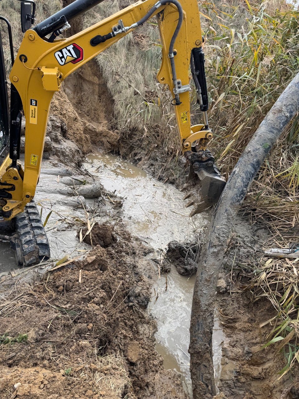 Żółta koparka CAT w błotnistym wykopie obok dużej rury, prawdopodobnie podczas prac instalacyjnych lub naprawczych. Trawa i roślinność w tle.