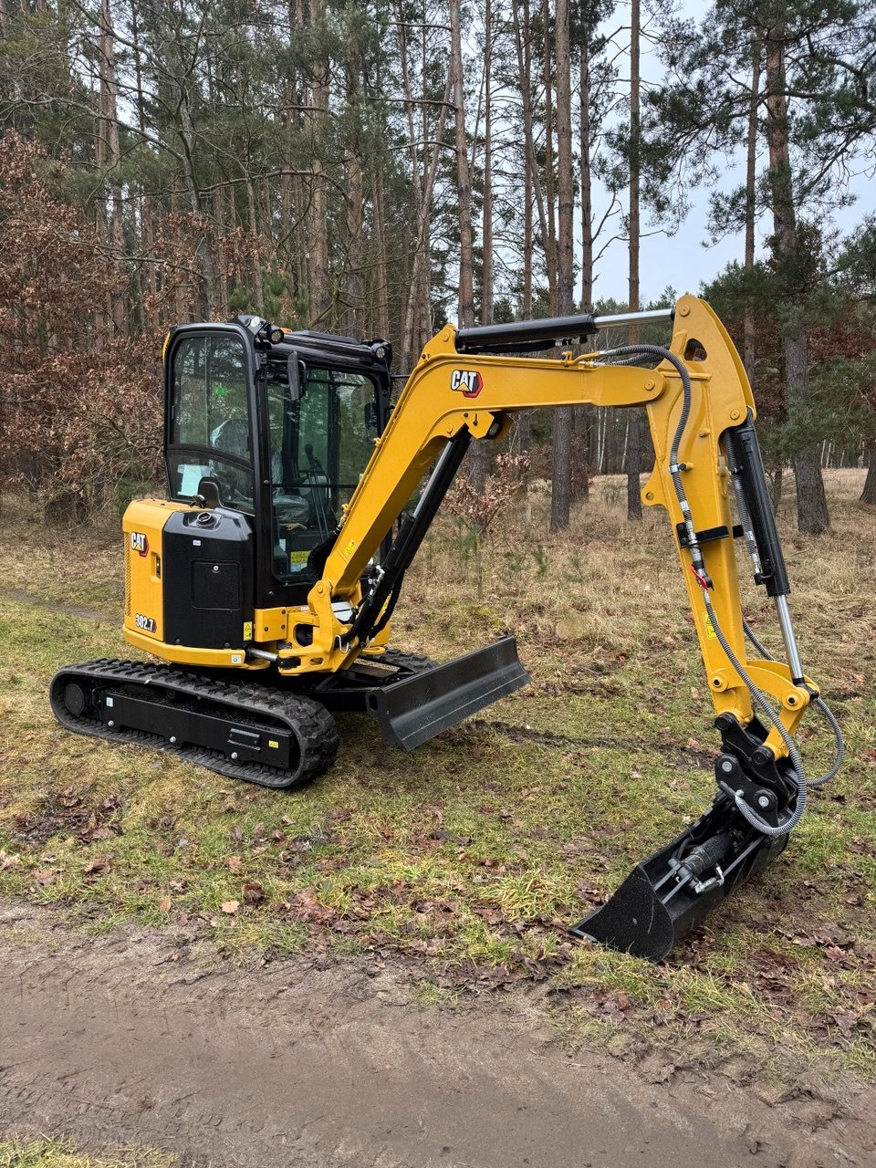 Żółto-czarna mini koparka CAT 302.7 na trawiastym terenie z lasem w tle. Łyżka koparki częściowo zagłębiona w ziemi, widoczne ślady pracy.