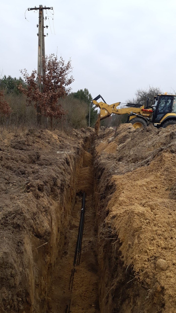 Głęboki wykop z rurami na dnie, zasypywany ziemią przez żółtą koparkę. W tle słup energetyczny i drzewa. Prace ziemne w toku, widok z góry.
