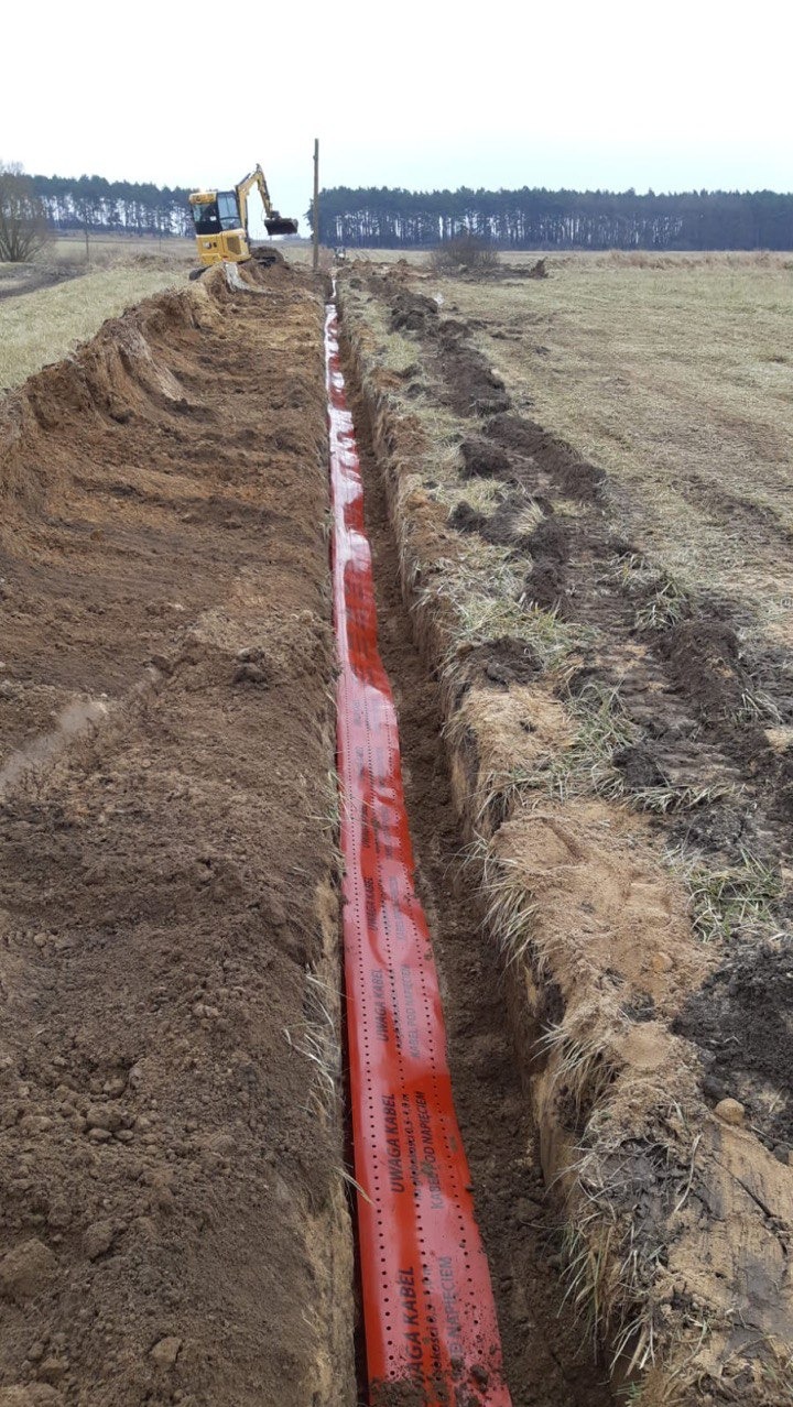 Długi wykop z czerwoną folią ostrzegawczą 'Uwaga kabel' w ziemi. W tle koparka i las.