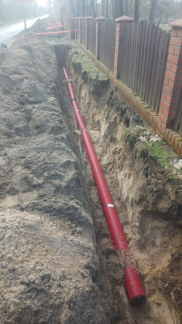Wykop z czerwoną rurą w ziemi, widoczny fragment płotu. Roboty ziemne w Krakowie. Rura karbowana ułożona w wykopie, przygotowana do zasypania.