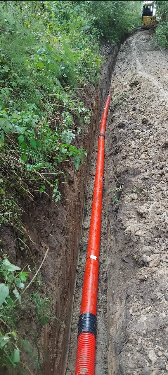 Czerwona rura w wykopie ziemnym, prawdopodobnie instalacja podziemna. W tle widoczna maszyna budowlana, a wokół bujna roślinność.