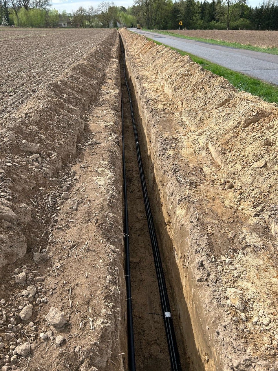 Długi wykop w polu z ułożonymi dwoma czarnymi rurami. Widok z góry na instalację w ziemi, obok asfaltowa droga i pole uprawne.