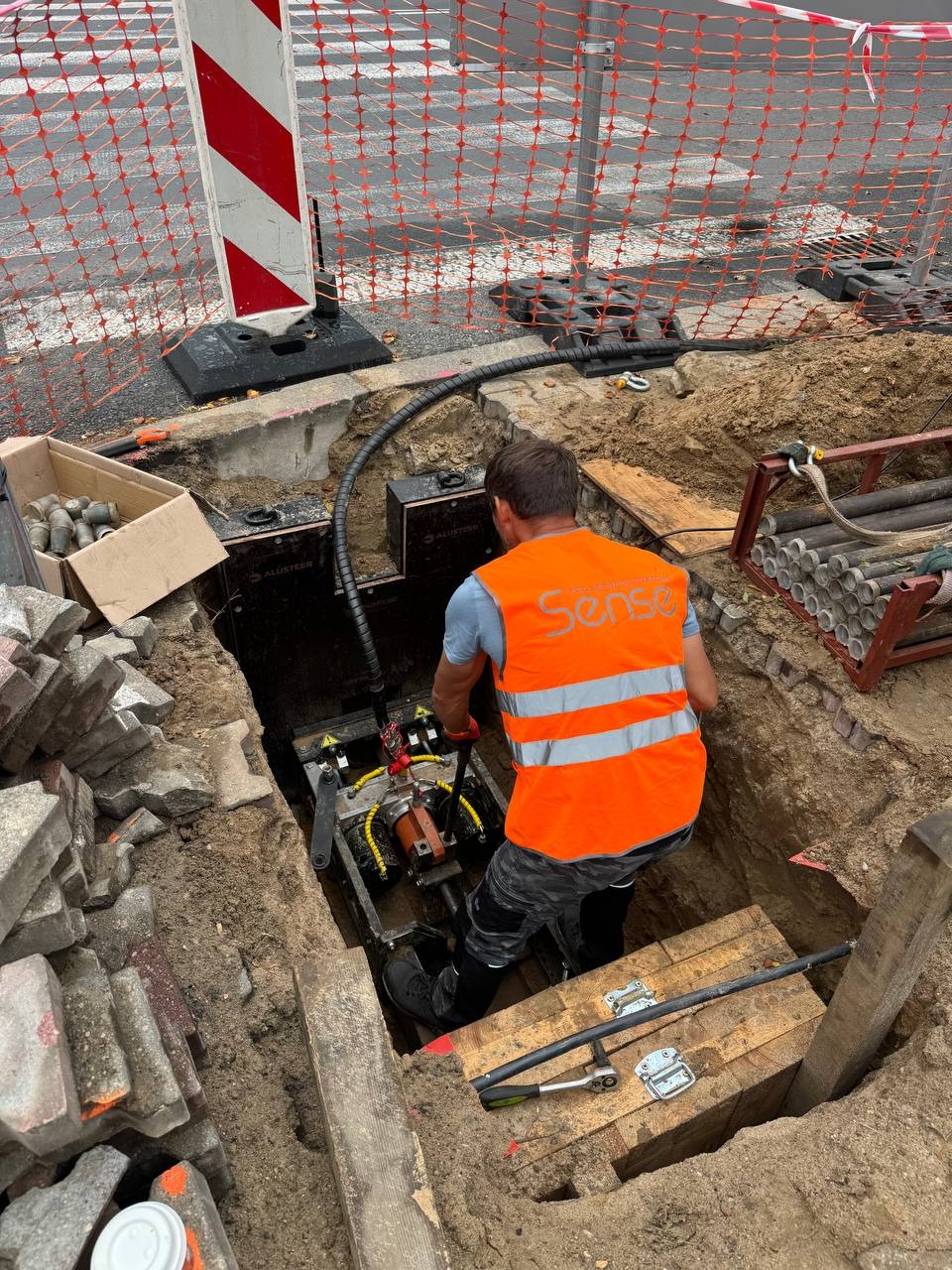 Pracownik w wykopie, w pomarańczowej kamizelce, obsługuje maszynę. Widoczne zabezpieczenia wykopu, kostka brukowa i elementy instalacji.