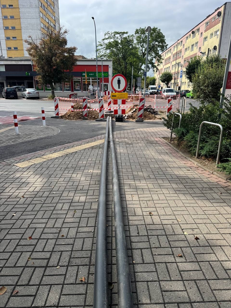 Oznakowany wykop na chodniku w Krakowie, zabezpieczony słupkami i siatką. Widoczne rury i znak zakazu przejścia. Prace ziemne w przestrzeni miejskiej.
