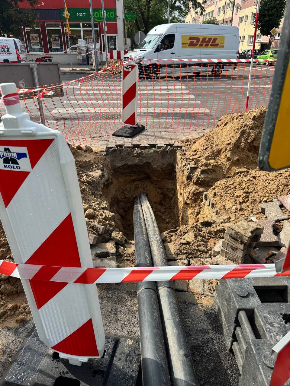 Wykop w chodniku z widocznymi rurami, zabezpieczony barierkami i taśmą ostrzegawczą. W tle budynki i przejeżdżający samochód dostawczy.