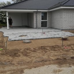 Masterwork - Układanie szarej kostki brukowej na piaskowym podłożu przed nowoczesnym domem z białej cegły, widoczne narzędzia i mężczyzna w tle.