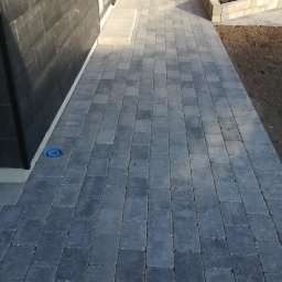 Masterwork - Ułożona, szara kostka brukowa prowadząca wzdłuż ściany budynku w Gdańsku, z widocznym włazem kanalizacyjnym i osobą pracującą w oddali przy rabacie kwiatowej.