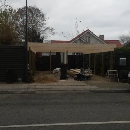 Masterwork - Konstrukcja drewnianej wiaty garażowej w trakcie budowy, widok z ulicy na szkielet z belek i słupów, narzędzia i materiały budowlane obok, w tle dom z czerwonym dachem.