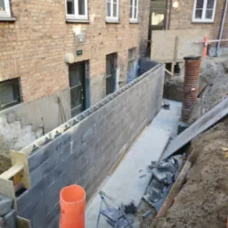 Wykop pod fundamenty budynku z widoczną ścianą fundamentową z szarych bloczków betonowych zbrojonych stalą, z deskowaniem szalunkowym na krawędzi,  rurą kanalizacyjną w wykopie i częściowo ułożoną...