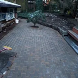 Ułożona kostka brukowa na podwórku z widocznym drzewkiem, fragmentem budynku z drewnianą werandą i stertą drewna opałowego w tle.