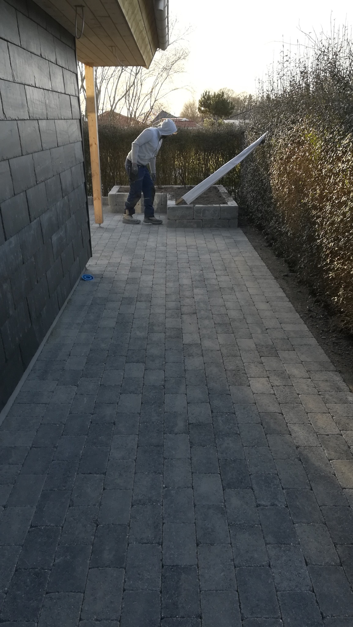Ułożona szara kostka brukowa na posesji, widoczny pracownik w bluzie z kapturem, przygotowujący obrzeże z kostki. W tle żywopłot i budynek z elewacją z ciemnego łupka.