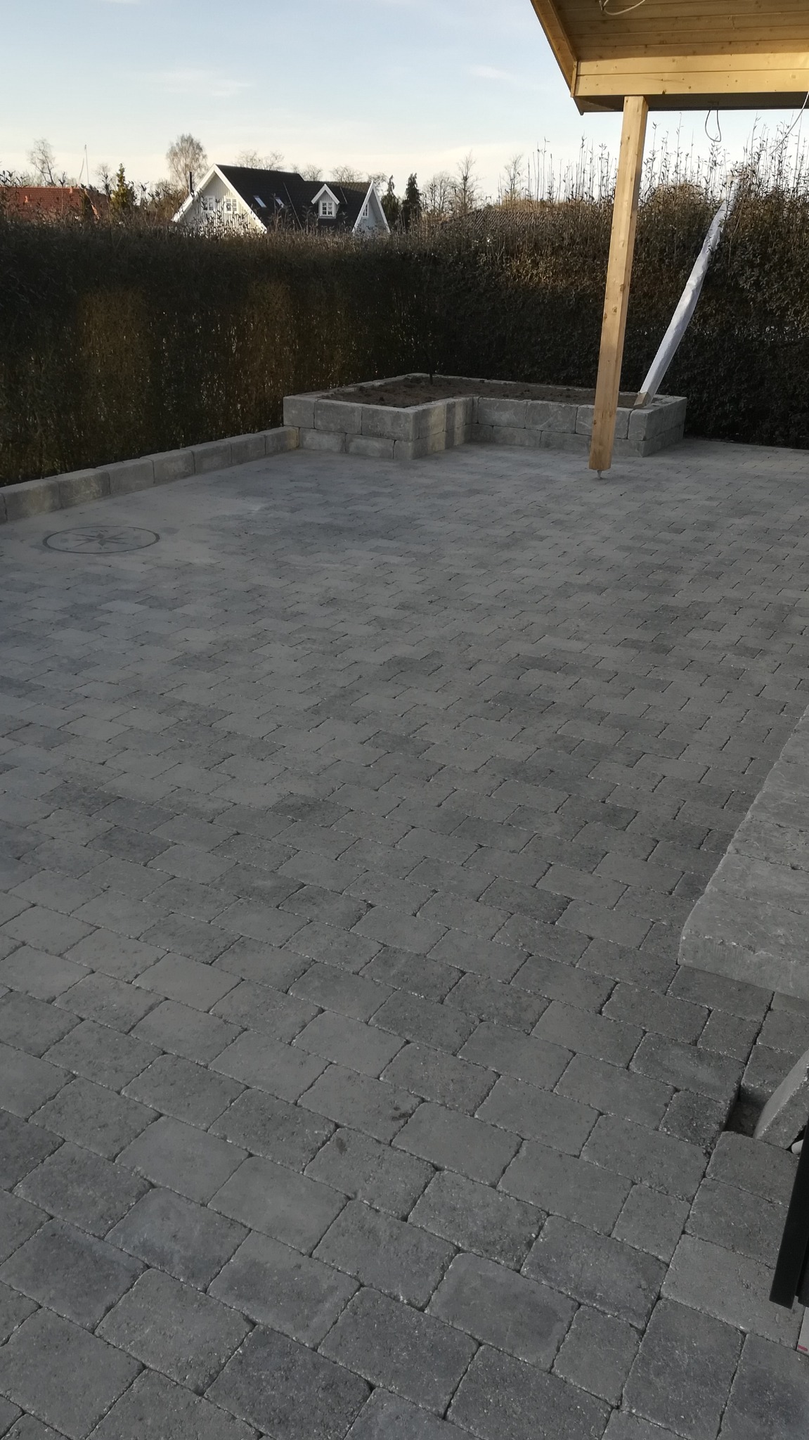Szara, prostokątna kostka brukowa ułożona na posesji, z widoczną studzienką i fragmentem drewnianej konstrukcji z słupem, w tle żywopłot i domy.