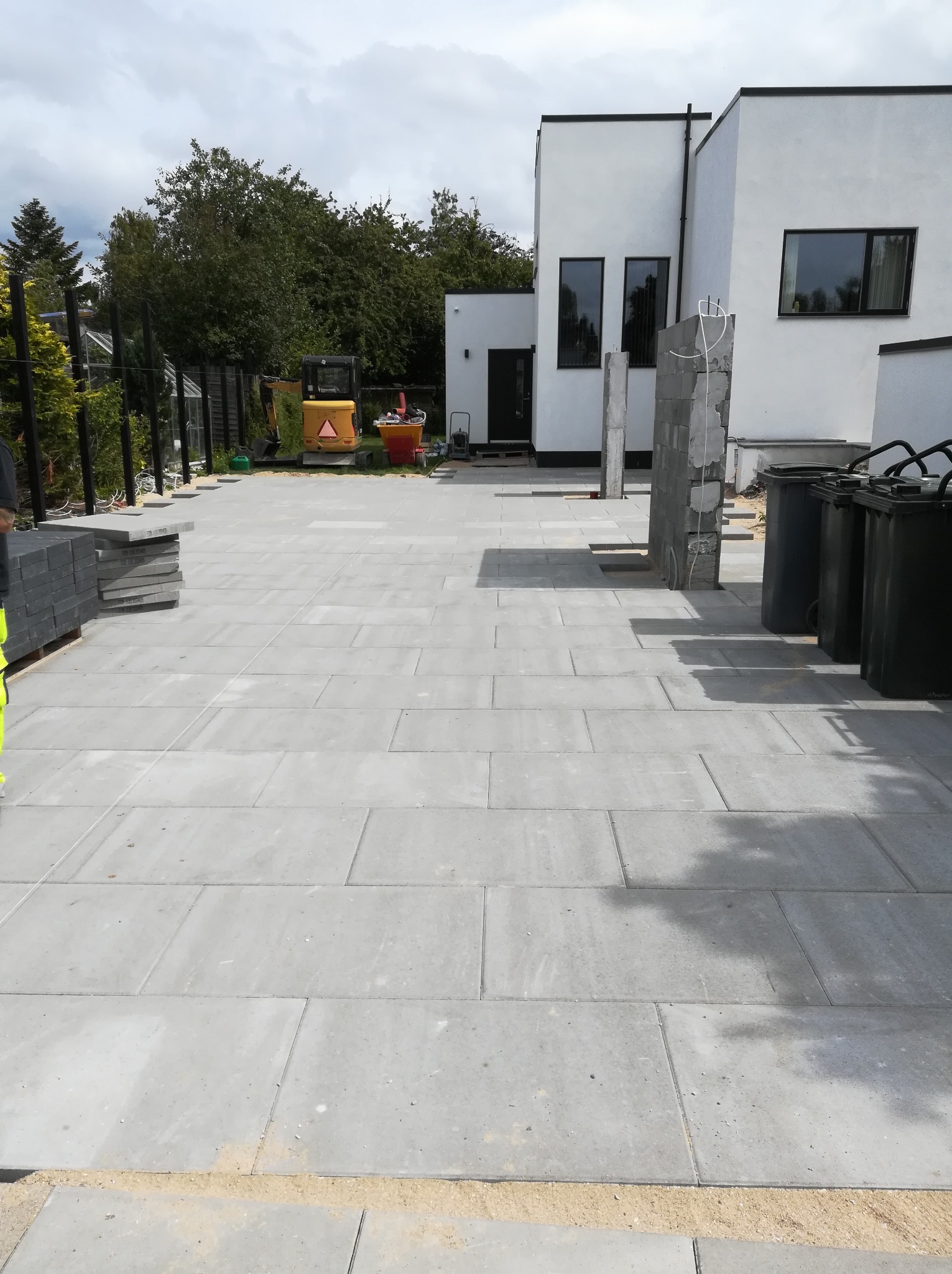 Ułożona szara kostka brukowa na posesji z widocznym minikoparką, paletą kostki, elementami fundamentów i pojemnikami na śmieci.