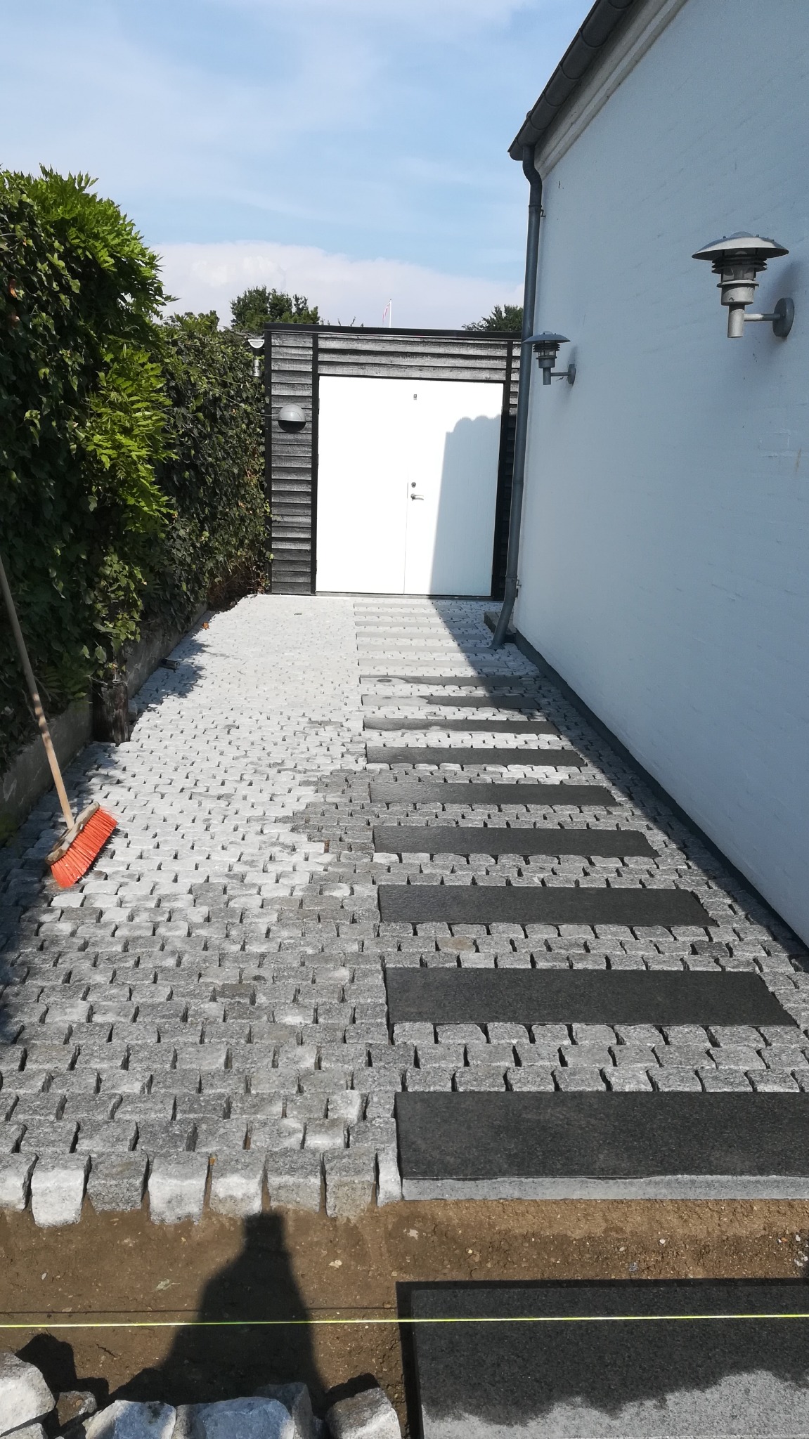Układanie szarej kostki brukowej z wkomponowanymi czarnymi płytami chodnikowymi, widoczne przygotowane podłoże i szczotka do zamiatania, w tle biały budynek i zielony żywopłot.