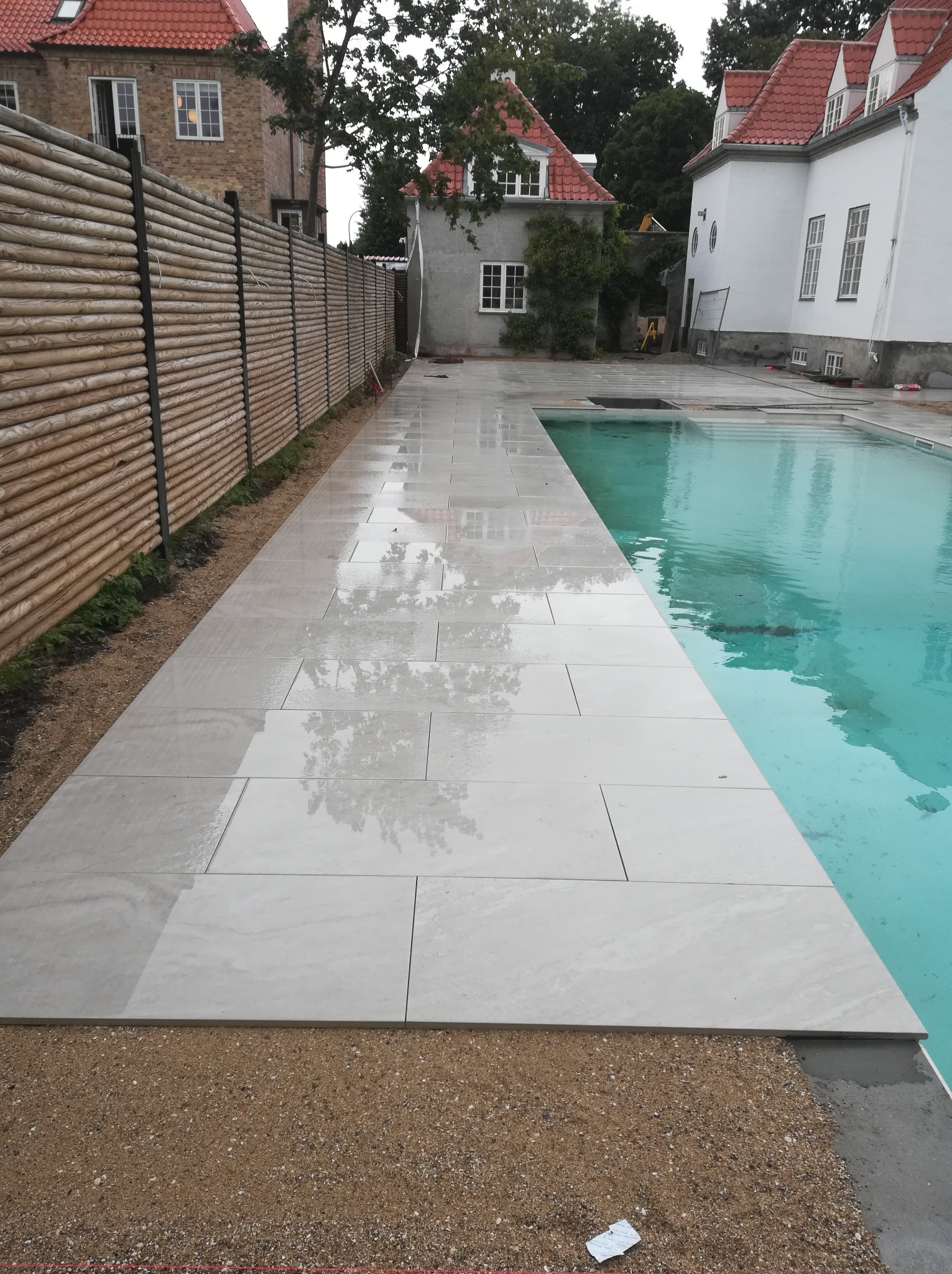 Jasne, prostokątne płyty ułożone wokół basenu, z widoczną drewnianą palisadą w tle i odbiciami budynków w mokrej powierzchni płyt.