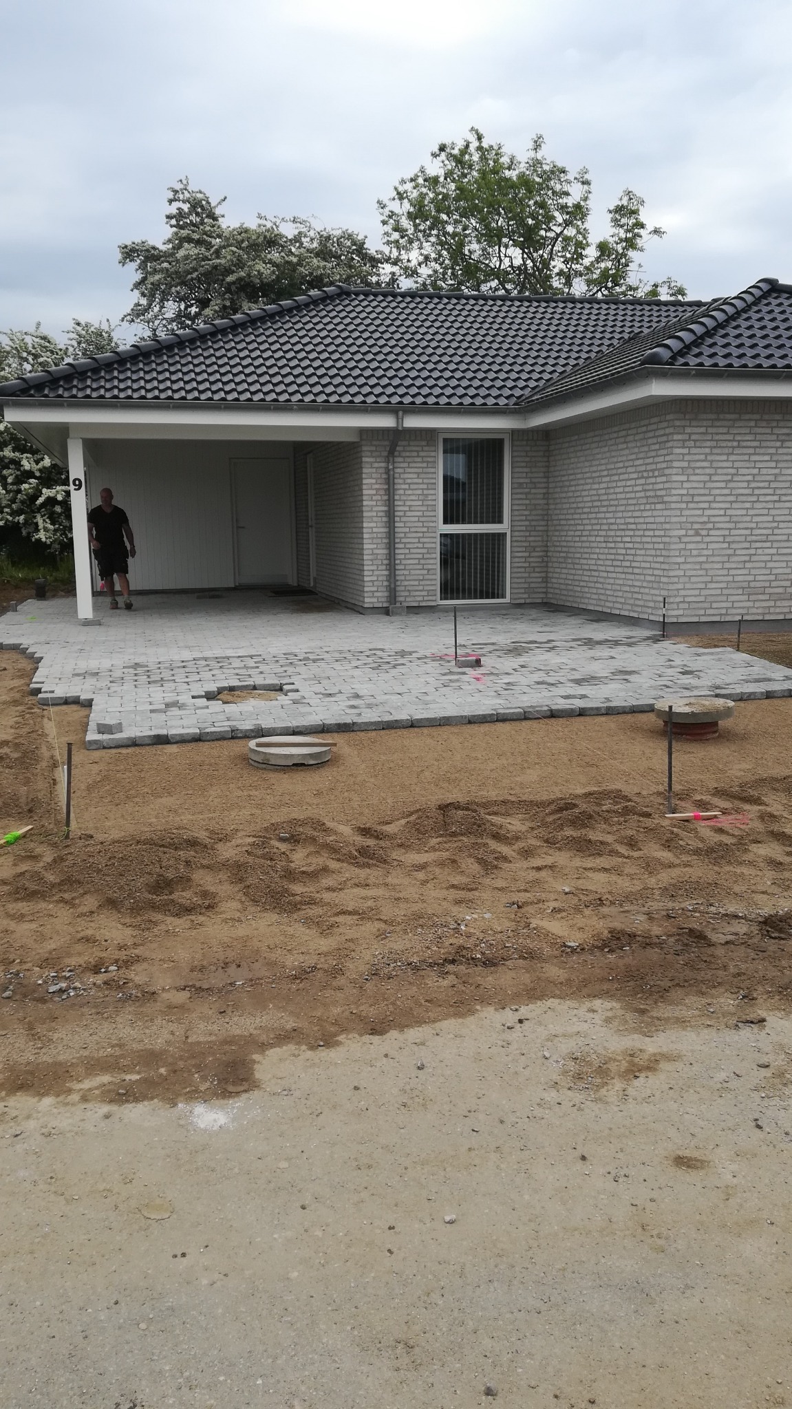 Układanie szarej kostki brukowej na piaskowym podłożu przed nowoczesnym domem z białej cegły, widoczne narzędzia i mężczyzna w tle.