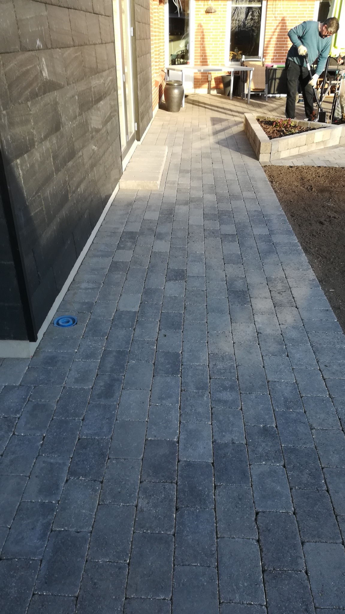 Ułożona, szara kostka brukowa prowadząca wzdłuż ściany budynku w Gdańsku, z widocznym włazem kanalizacyjnym i osobą pracującą w oddali przy rabacie kwiatowej.