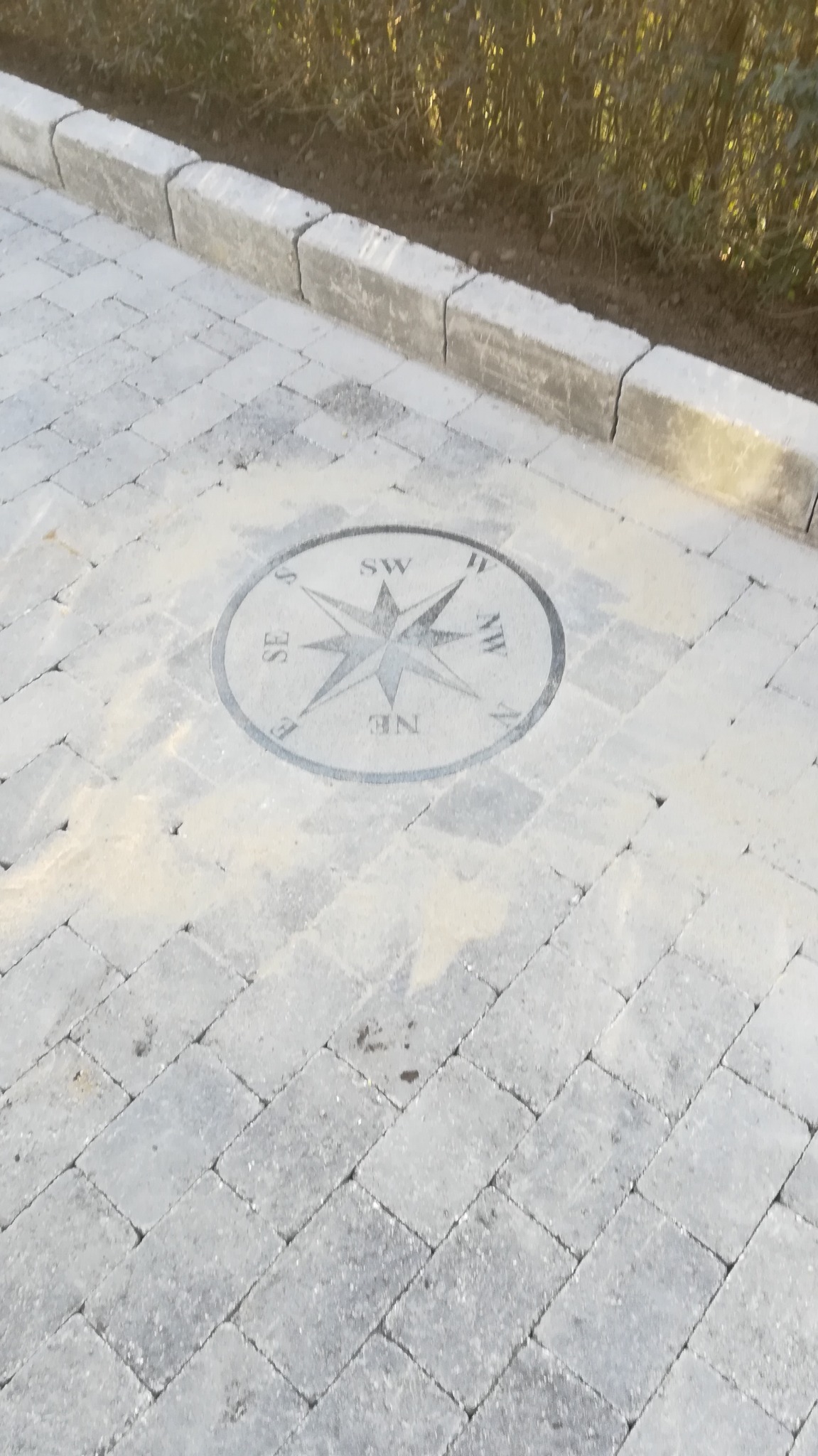 Szara kostka brukowa ułożona w jodełkę z dekoracyjnym elementem w kształcie kompasu pośrodku, obrzeża z szarych kostek.