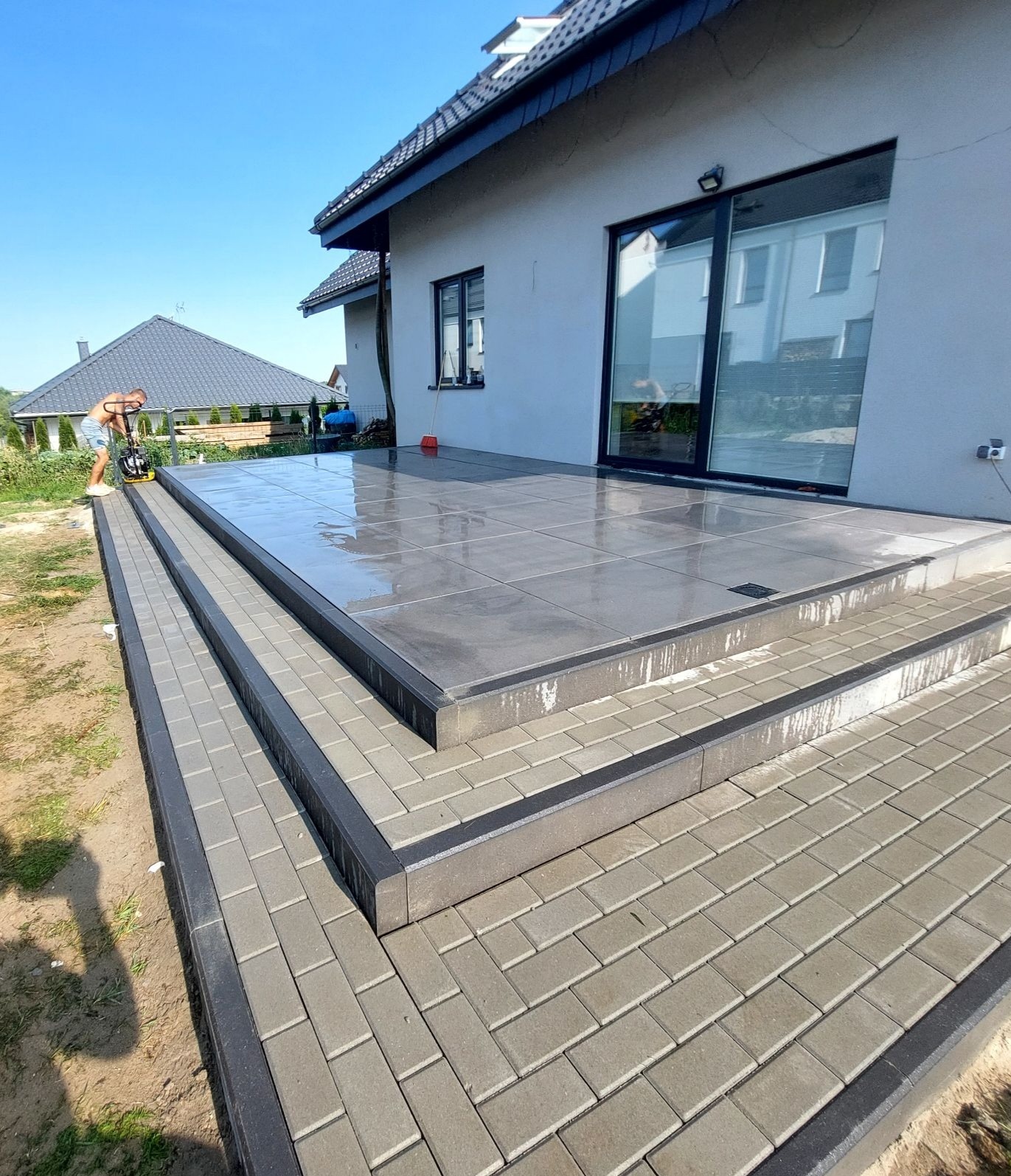 Mężczyzna zagęszcza wibratorem płytowym świeżo ułożoną, mokrą nawierzchnię z płyt betonowych na tarasie z kostki brukowej przy domu.