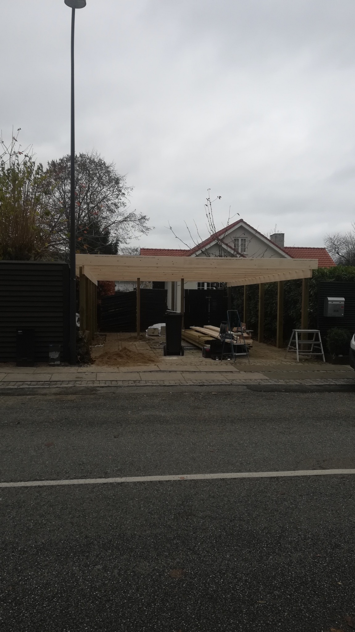 Konstrukcja drewnianej wiaty garażowej w trakcie budowy, widok z ulicy na szkielet z belek i słupów, narzędzia i materiały budowlane obok, w tle dom z czerwonym dachem.