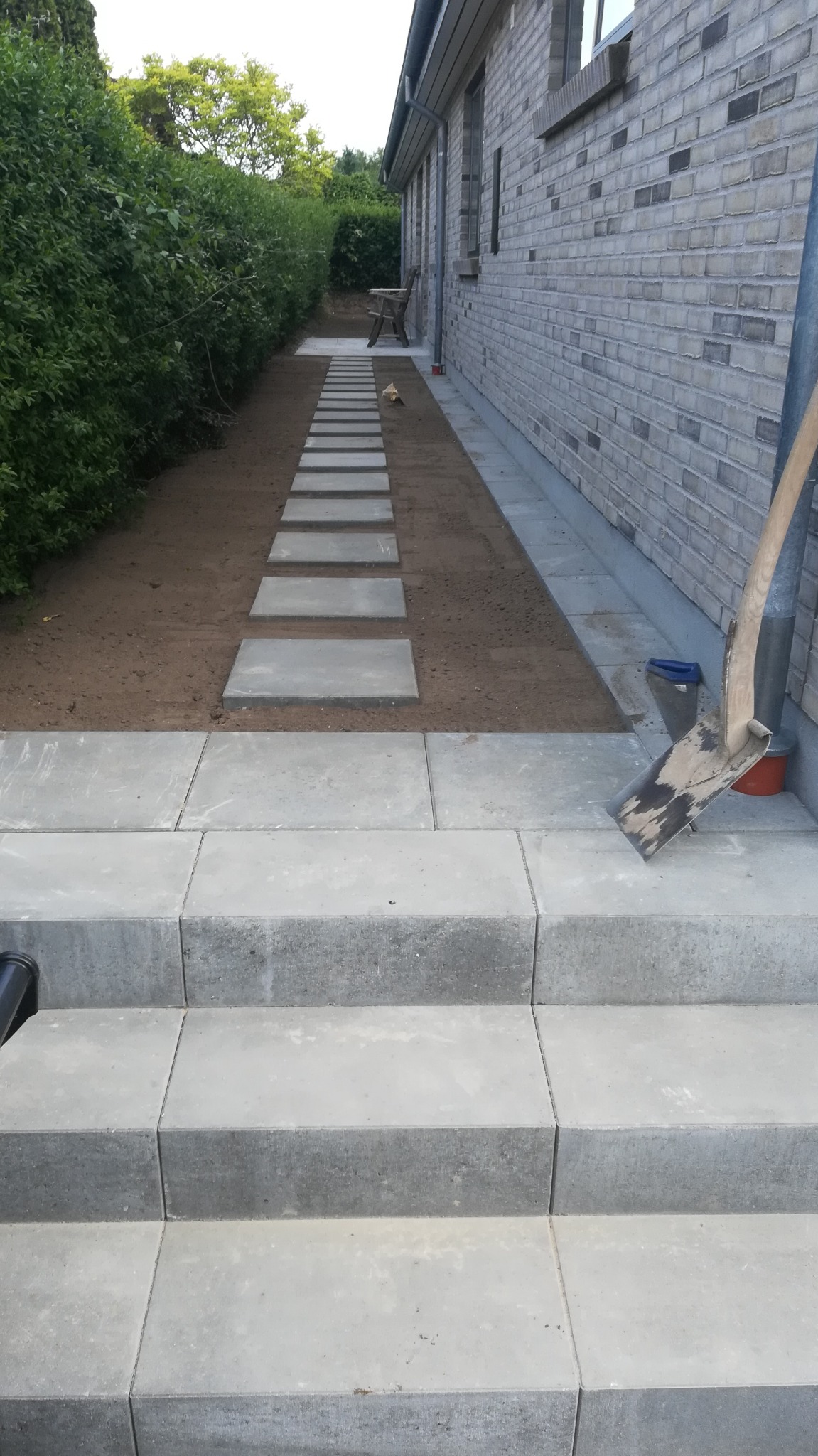 Stopnie z kostki brukowej prowadzące do ścieżki z płyt, widok z dołu, obok ściana budynku z jasnej cegły i zielony żywopłot, w tle drzewa.