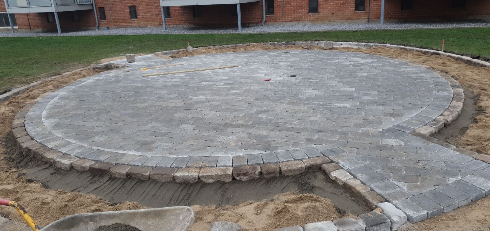Okazały, okrągły plac z szarej kostki brukowej w trakcie budowy, otoczony obrzeżem z kamieni, z widoczną żółtą taczką i piaskiem.