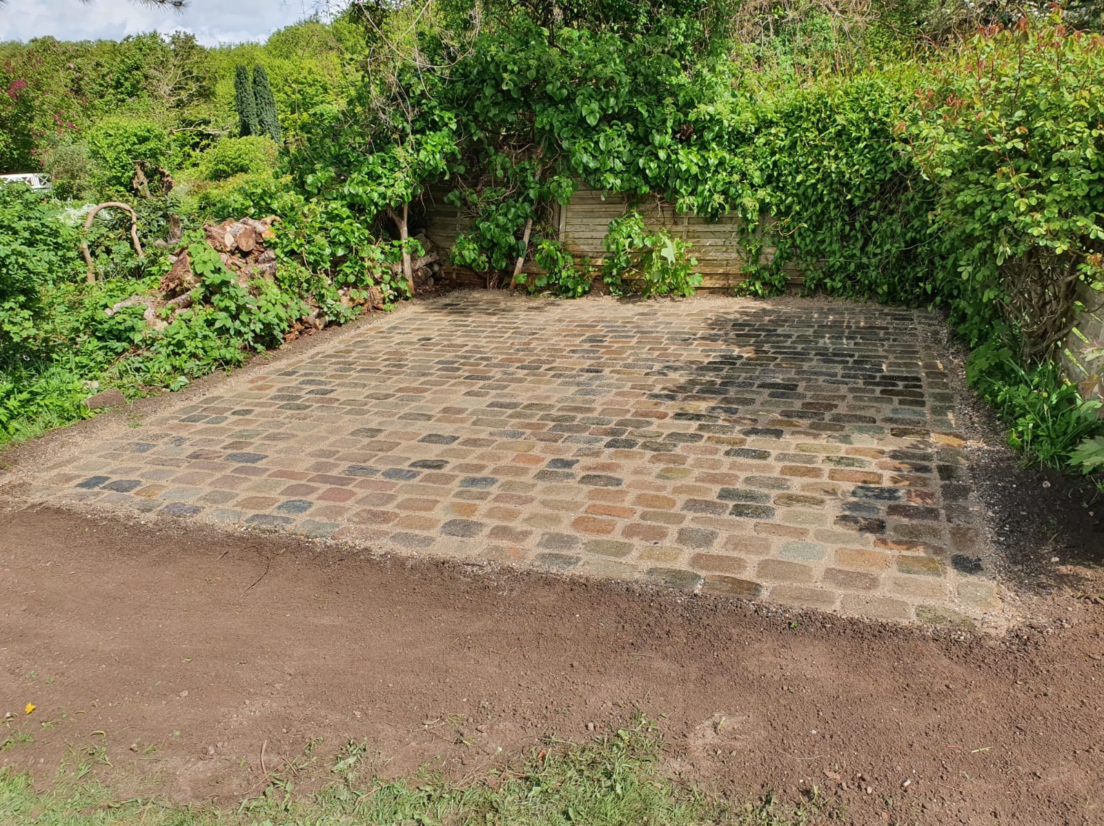 Równa powierzchnia z kostki brukowej w odcieniach brązu i szarości, otoczona przygotowanym podłożem z ziemi, z drewnianym płotem i bujną zielenią w tle.