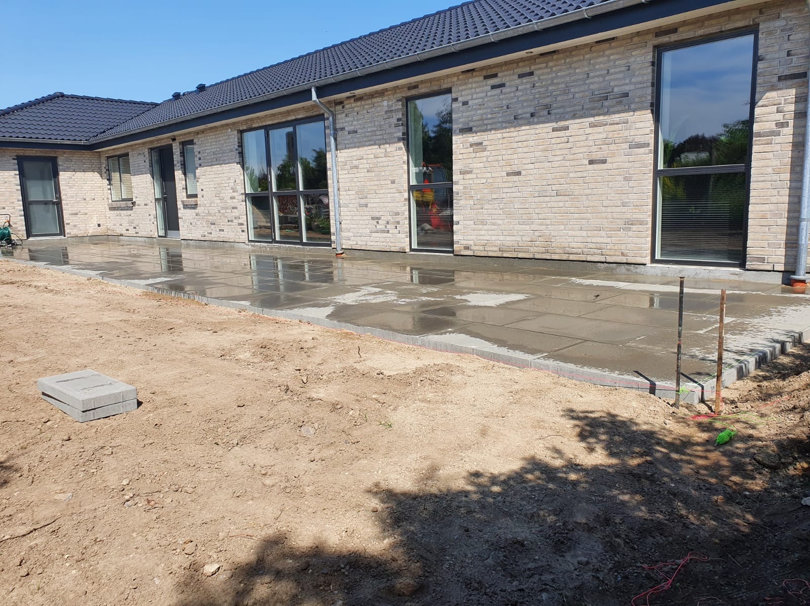 Świeżo ułożona, mokra nawierzchnia z dużych, szarych płyt betonowych przylegająca do elewacji domu z cegły klinkierowej; widoczne przygotowane elementy obrzeża i niezabezpieczony grunt.