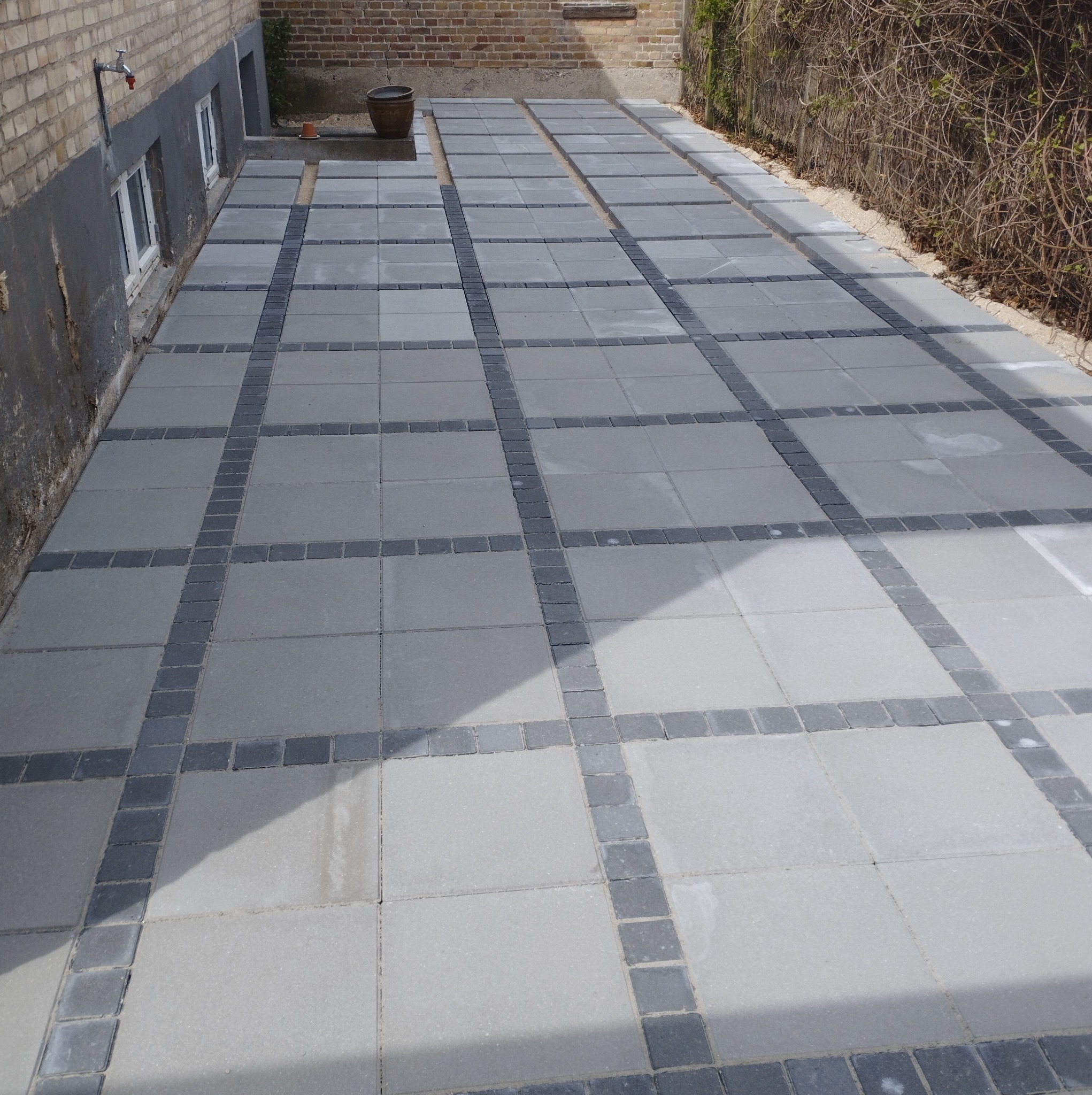 Świeżo ułożony chodnik z jasnoszarej kostki brukowej z ciemnoszarymi obwódkami, biegnący wzdłuż ściany budynku z cegły