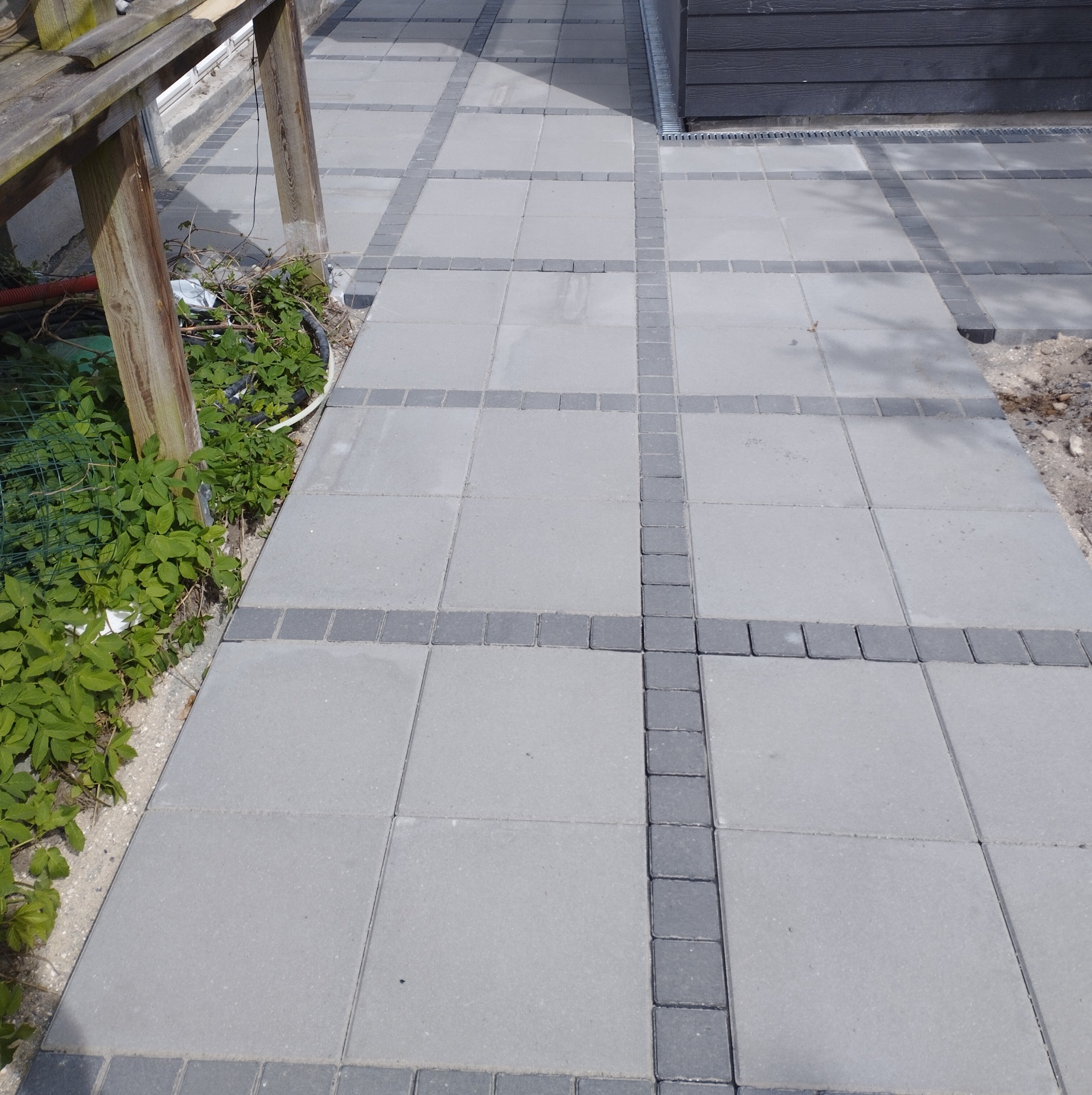 Ułożona szara kostka brukowa z ciemnoszarą obwódką, widoczny fragment drewnianej konstrukcji i zieleni