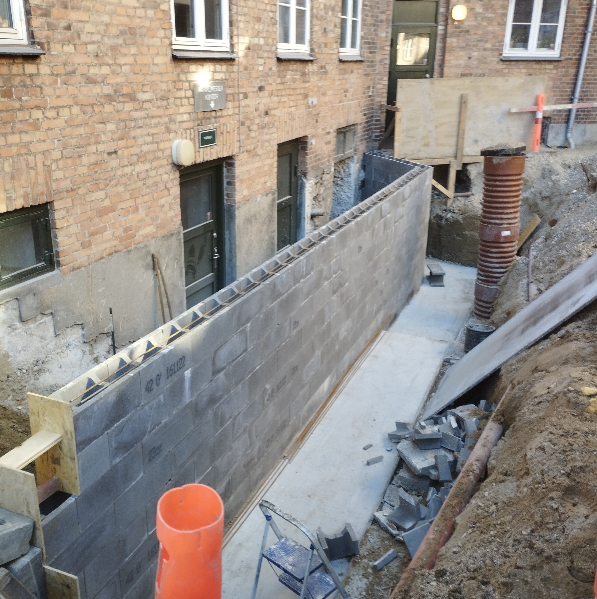 Wykop pod fundamenty budynku z widoczną ścianą fundamentową z szarych bloczków betonowych zbrojonych stalą, z deskowaniem szalunkowym na krawędzi,  rurą kanalizacyjną w wykopie i częściowo ułożoną...