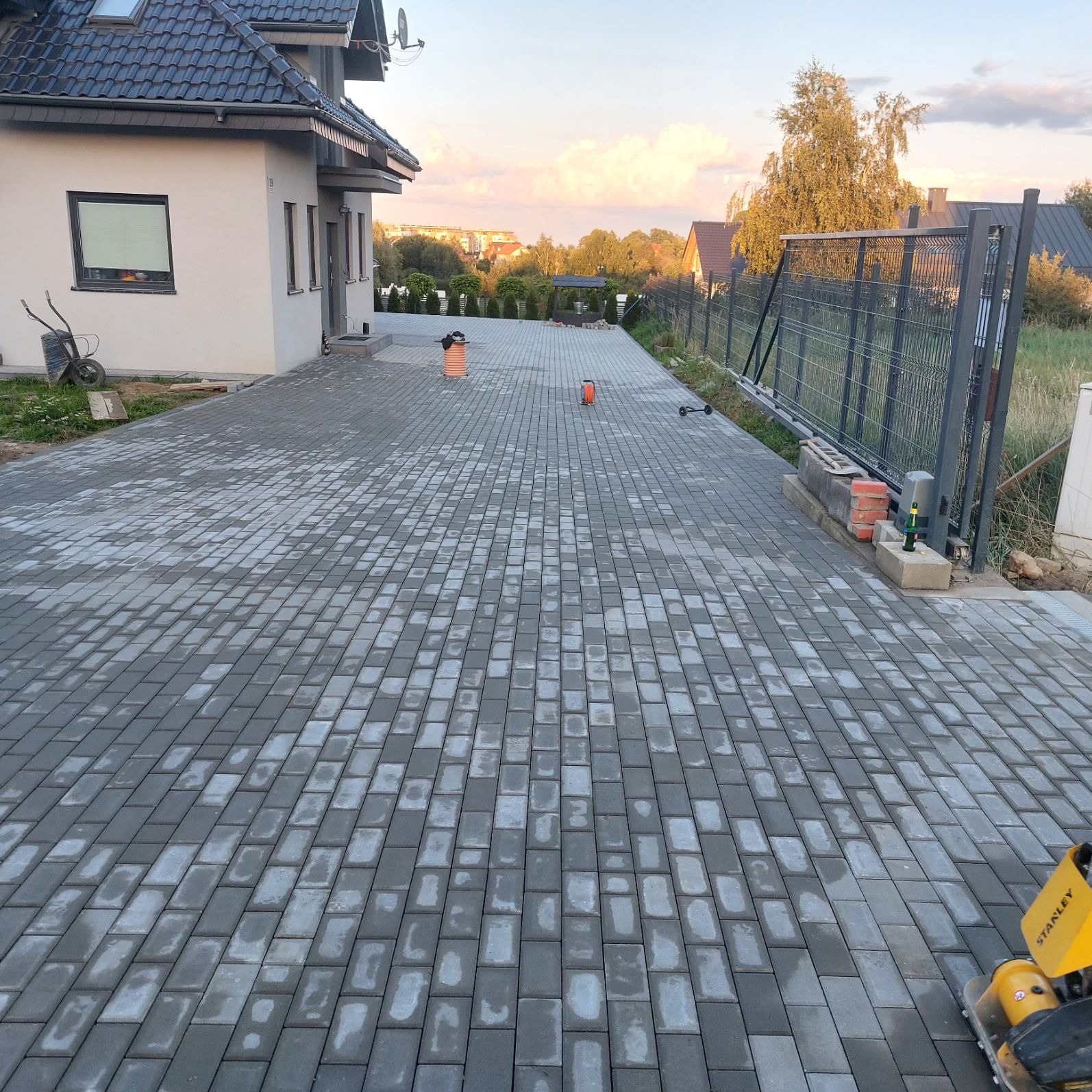 Ułożona szara kostka brukowa na podjeździe do domu jednorodzinnego z bramą przesuwną, widoczna ubijarka w prawym dolnym rogu.