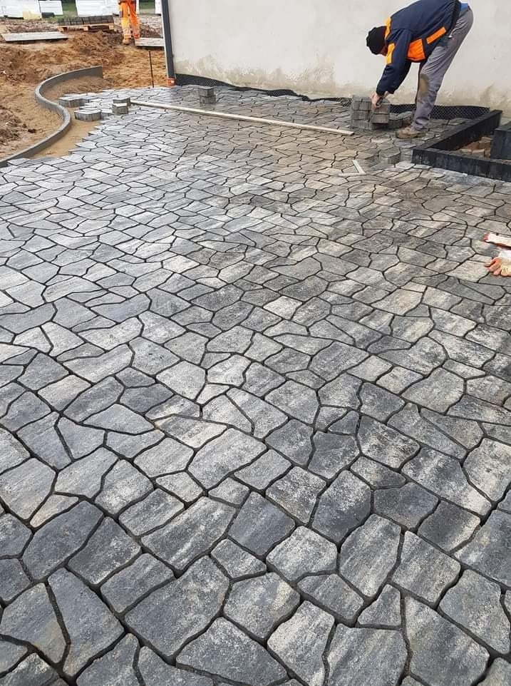 Układanie nieregularnej, grafitowej kostki brukowej na posesji, widoczny pracownik w trakcie montażu obrzeży i wyrównywania podłoża.