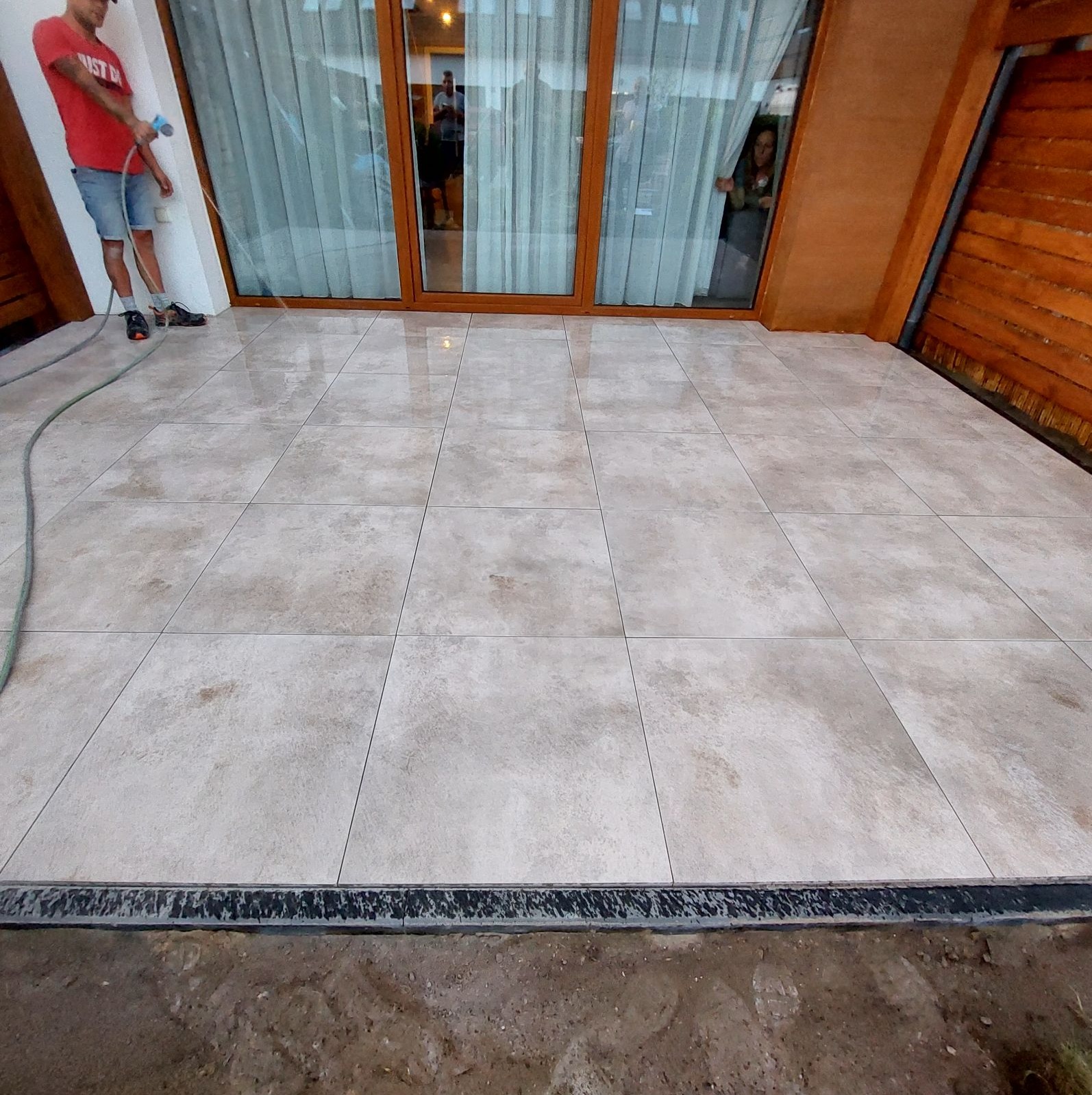 Jasne, beżowe płytki podłogowe na tarasie, czyszczone wodą pod ciśnieniem przez mężczyznę w czerwonej koszulce, widoczny drenaż liniowy przy krawędzi, z przeszkleniami budynku w tle.