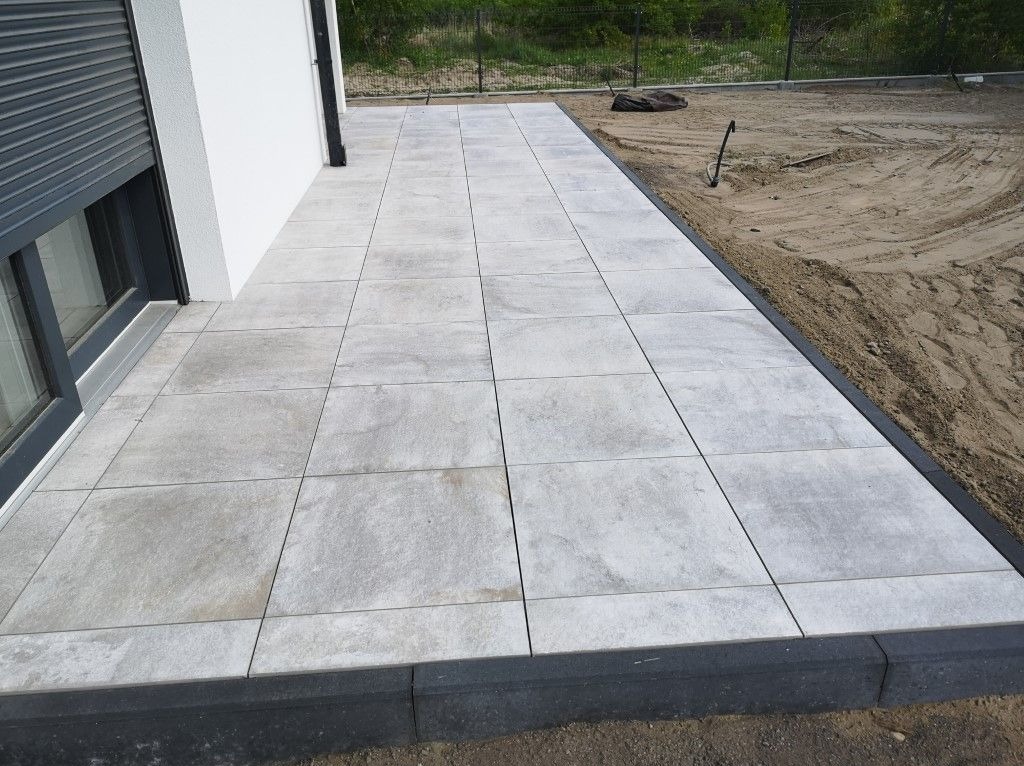 Jasnoszara, kwadratowa nawierzchnia z płytek ułożona przy ścianie budynku z roletami okiennymi, obrzeże z ciemnych kostek, obok widoczny piasek.