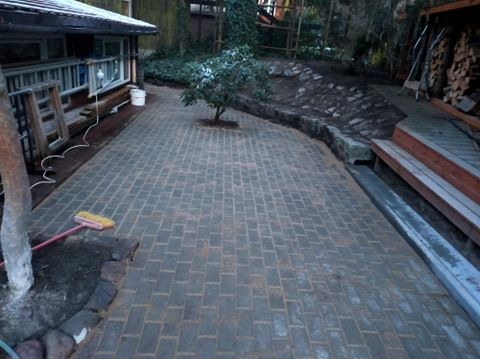 Ułożona kostka brukowa na podwórku z widocznym drzewkiem, fragmentem budynku z drewnianą werandą i stertą drewna opałowego w tle.