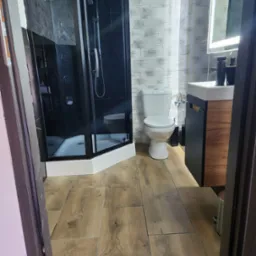 Nowoczesna łazienka z czarną kabiną prysznicową typu walk-in, drewnopodobnymi płytkami podłogowymi, toaletą i umywalką z szafką w odcieniach czerni i drewna.