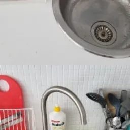 Metalowy zlew kuchenny z widocznym odpływem i śladami użytkowania, obok bateria, płyn do naczyń, gąbka, ociekacz na naczynia i przybory kuchenne na tle białej ściany z drobną mozaiką.