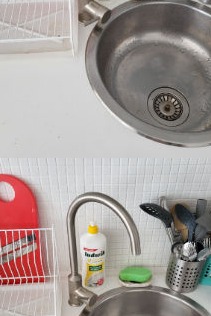 Metalowy zlew kuchenny z widocznym odpływem i śladami użytkowania, obok bateria, płyn do naczyń, gąbka, ociekacz na naczynia i przybory kuchenne na tle białej ściany z drobną mozaiką.