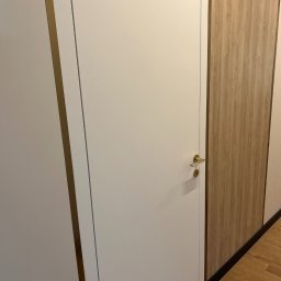 Artur Wolak Bud - Białe drzwi wewnętrzne z delikatną złotą klamką i ramą, sąsiadujące z panelem ściennym o wzorze jasnego drewna, uzupełnione brązową listwą przypodłogową.