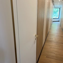 Artur Wolak Bud - Długi korytarz z drewnianą podłogą i szeregiem białych drzwi z złotymi klamkami, przechodzący w jasne, przeszklone zakończenie z widokiem na zieleń.