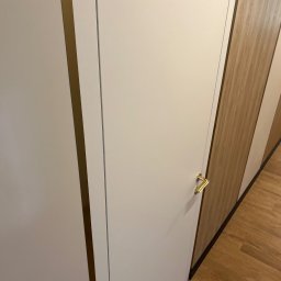 Artur Wolak Bud - Białe, minimalistyczne drzwi wewnętrzne z złotą klamką, widoczne na tle drewnianej podłogi i zabudowy meblowej z elementami w kolorze drewna.