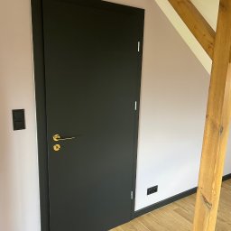 Artur Wolak Bud - Czarna, matowa, bezprzylgowa drzwi z złotą klamką na tle jasnoróżowej ściany z drewnianą belką stropową i czarnymi listwami przypodłogowymi oraz gniazdkami, widoczny klimatyzator.