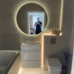 Artur Wolak Bud - Nowoczesna łazienka z podświetlanym lustrem w kształcie koła i białymi płytkami na ścianach, widoczna umywalka z szafką, toaleta i wanna, z odbiciem osoby robiącej zdjęcie w lustrze.